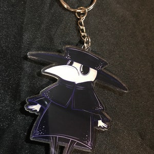 Pu&ograve; includere: Un portachiavi a forma di medico della peste nero e bianco con un lungo becco e un cappello nero. Il portachiavi &egrave; in acrilico trasparente e ha una catena argentata.