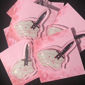 May include: A set of four clear acrylic pins featuring a white snake wrapped around a black knife. The pins are on a pink background with a watercolor effect.