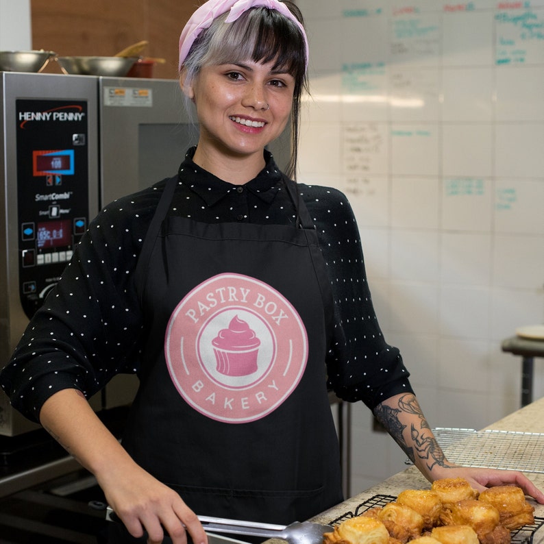 May include: A woman wearing a black apron with a pink logo that says "Pastry Box Bakery" and a pink cupcake graphic. She is standing in a kitchen and holding a metal spatula.