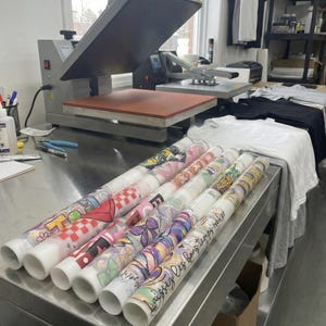 May include: Several rolls of heat transfer vinyl with various colorful graphic designs, including checkered patterns, floral motifs, and the text Laissez les bons temps rouler, arranged on a stainless steel table near a heat press machine.
