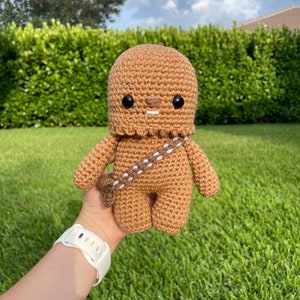 Op de afbeelding: Een bruin gehaakt Chewbacca pluche speelgoed met zwarte ogen en een witte bandolier. Het speelgoed wordt in de hand van een persoon gehouden.