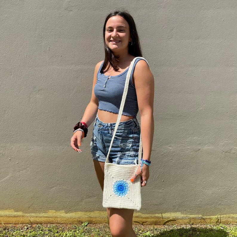 May include: A white crocheted crossbody bag with a blue and white flower design. The bag is being worn by a person wearing a blue and white striped tank top and denim shorts.