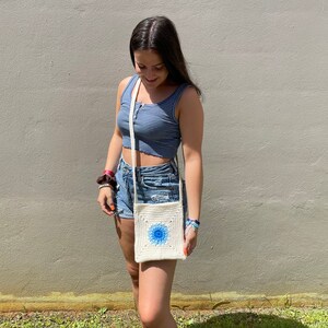 May include: A woman wearing a blue and white striped tank top and denim shorts is carrying a white crocheted crossbody bag with a blue floral design.