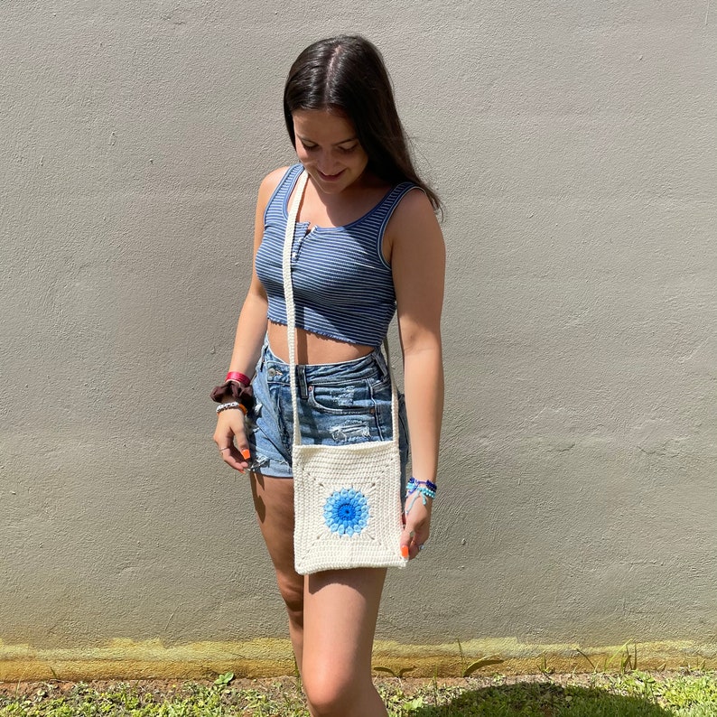 May include: A woman wearing a blue and white striped tank top and denim shorts is carrying a white crocheted crossbody bag with a blue floral design.