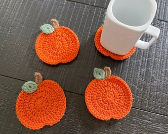Puede incluir: Cuatro posavasos de ganchillo naranja hechos a mano con hojas verdes y tallos marrones. Una taza blanca cuadrada descansa sobre uno de los posavasos. Los posavasos están sobre una superficie de madera oscura.