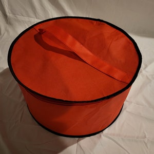 May include: A red circular storage bag with a black zipper and a black handle. The bag is made of a soft, durable fabric.