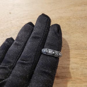 May include: A silver ring with an openwork design featuring small, round gemstones. The ring is displayed on a black surface, showcasing its intricate details and craftsmanship. The ring's design includes a series of diamond-shaped patterns.