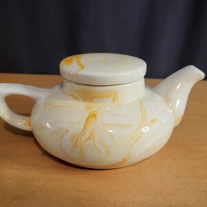 May include: Cream-colored ceramic teapot with a lid, featuring a swirled yellow pattern. The teapot has a curved handle and a spout. The design is reminiscent of marble. The teapot is sitting on a wooden surface.