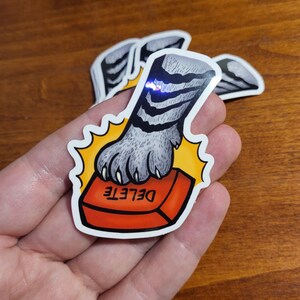 May include: A sticker featuring a cat paw pressing a red "DELETE" button. The paw is gray with black stripes, and the button is outlined in yellow. Several stickers are visible.