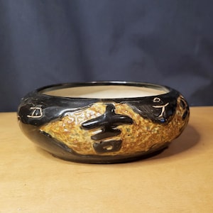 May include: A black and tan ceramic bowl with a textured exterior and a smooth interior. The bowl features embossed Asian characters and a glossy black rim. The bowl is suitable for decorative or functional use.