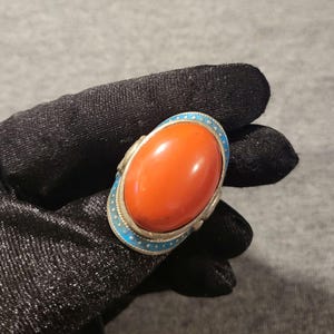 May include: A ring featuring a large, smooth, oval-shaped orange stone set in a silver-toned metal frame. The setting is accented with a border of bright blue enamel featuring small white dots, creating a distinct contrast against the orange center.