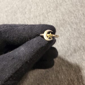 May include: A silver ring featuring a crescent moon and a star design. The moon is white, and the star is gold. The ring is held by a black glove against a gray background. The ring is a celestial-themed jewelry item.