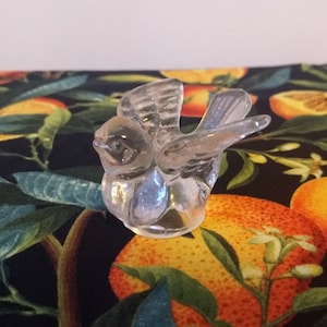 May include: A clear glass bird figurine with a detailed wingspan and a small beak. The figurine is sitting on a black background with a floral pattern.