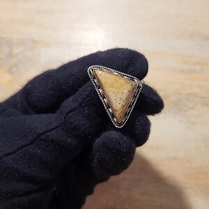 May include: A triangular ring with a speckled, tan-colored stone set in a silver-toned metal frame. The frame has a twisted rope design around the stone. The ring is held by a gloved hand.