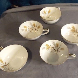 May include: Five white teacups with gold trim and handles. Each cup features a wheat stalk design in gold and gray. The cups are arranged on a gray surface, showcasing a classic and elegant aesthetic.