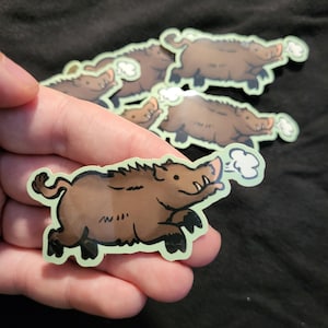 May include: A collection of cartoon-style stickers featuring a brown boar with a light green outline. The boar is depicted running, with steam or breath emanating from its snout. The stickers are held in a hand against a dark background.