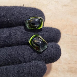 May include: Two glass rings with a dark center and a green and yellow border. The rings are rectangular in shape and appear to be handmade. The rings are displayed on a dark blue surface.