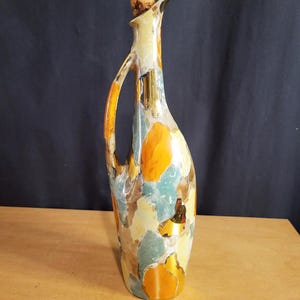 May include: A tall, decorative ceramic bottle with a cork stopper. The bottle features a handle and spout, and is painted with a colorful abstract design in shades of orange, yellow, blue, and brown. The bottle is approximately 12 inches tall.
