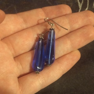 May include: A pair of blue teardrop earrings. The earrings have a long, slender shape with a rounded bottom and a silver-colored hook. The blue glass has a glossy finish and is accented with small silver beads.