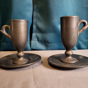 May include: Two silver-colored cups with matching saucers. The cups have a rounded, slightly flared shape with a handle on one side. The saucers are round and have a slightly raised edge.
