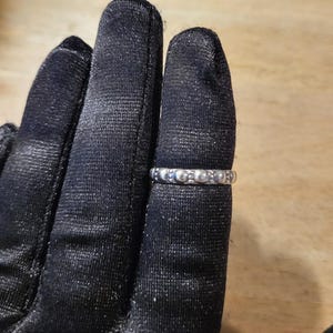 May include: A silver ring with a repeating pattern of small, raised circular designs. The ring is shown on a black glove, with a wooden surface in the background. The ring appears to be made of a shiny metal.