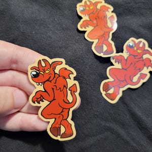 May include: Three red devil stickers with a tan border. Each sticker features a cartoon devil with horns, a tail, and a mischievous expression. The stickers are on a black background.