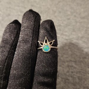 May include: A silver-toned ring featuring a circular, iridescent blue-green stone set in the center. The band includes a decorative crown-like element with three pointed, open-work arches extending upward from the base of the stone setting.