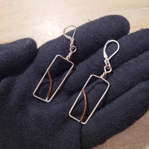 May include: A pair of silver-tone rectangular earrings with a copper-colored accent. The earrings have a lever-back closure and are displayed on a black surface.