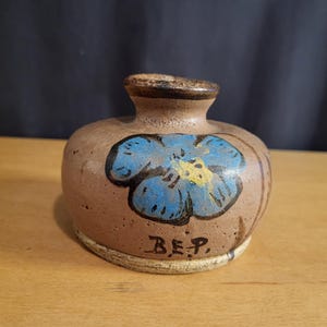 May include: A small, brown ceramic vase with a blue flower painted on the side. The vase has a white rim and the initials "B.E.P." are painted on the bottom.