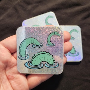 May include: Square holographic stickers featuring a teal sea monster design. The sea monsters are depicted emerging from the water in various poses. The stickers have a glittery, iridescent background and are held in a hand.