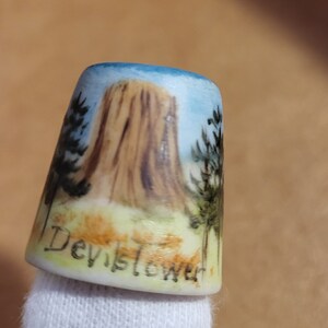 May include: A white ceramic thimble with a hand-painted image of Devil's Tower National Monument in Wyoming. The thimble has a blue sky, brown rock formation, and green trees. The words "Devil's Tower" are written on the bottom of the thimble.