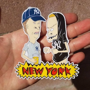 May include: Two cartoon characters, one wearing a blue baseball cap with a New York Yankees logo and a striped shirt, the other wearing a black hoodie with a New York Yankees logo, stand in front of a yellow banner with the words "NEW YORK" in red letters.