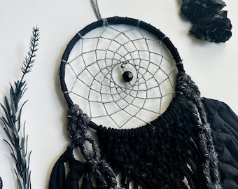 May include: A black and gray dreamcatcher with a woven web design and a central black bead. The dreamcatcher features black fabric accents and hanging strands. A sprig of dark foliage and black paper roses are also visible.