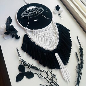 May include: A black and white macrame wall hanging featuring an embroidered UFO design. The circular embroidery hoop has a black background with a white UFO and a figure being abducted. The macrame has white and black fringe.