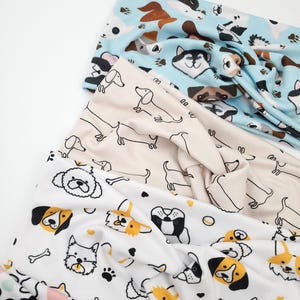 May include: Three fabric headbands with dog-themed patterns. One is light blue with various dog breeds, another is beige with dachshunds, and the third is white with dog faces, paw prints, and bones. The headbands are knotted at the top.