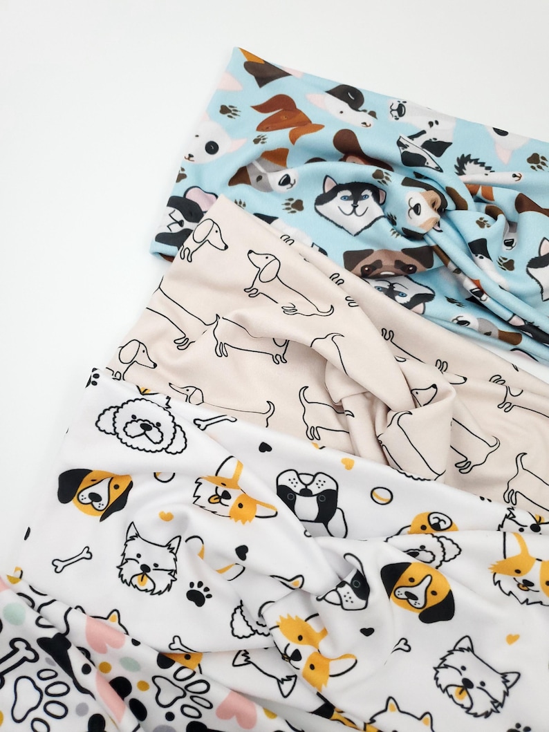 May include: Three fabric headbands with dog-themed patterns. One is light blue with various dog breeds, another is beige with dachshunds, and the third is white with dog faces, paw prints, and bones. The headbands are knotted at the top.