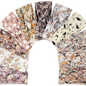 May include: A collection of ten fabric headbands arranged in a fan shape, featuring various patterns including ghosts, flowers, black cats, stars, moons, pumpkins, rainbows, and text reading Trick or Treat and Hocus Pocus in muted seasonal colors.