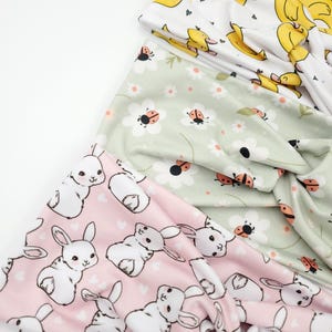 May include: A stack of fabric headbands with various patterns. One headband features yellow ducks on a white background. Another has white bunnies on a pink background. A third headband displays ladybugs and daisies on a light green background.