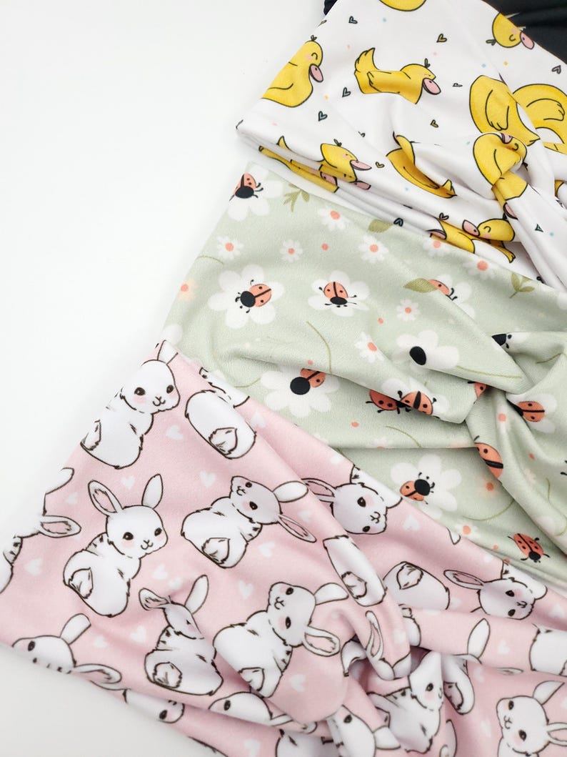 May include: A stack of fabric headbands with various patterns. One headband features yellow ducks on a white background. Another has white bunnies on a pink background. A third headband displays ladybugs and daisies on a light green background.