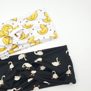 May include: Two headbands with a twist design. One is white with yellow duck illustrations and small hearts. The other is black with white goose illustrations. Both are made of soft fabric.