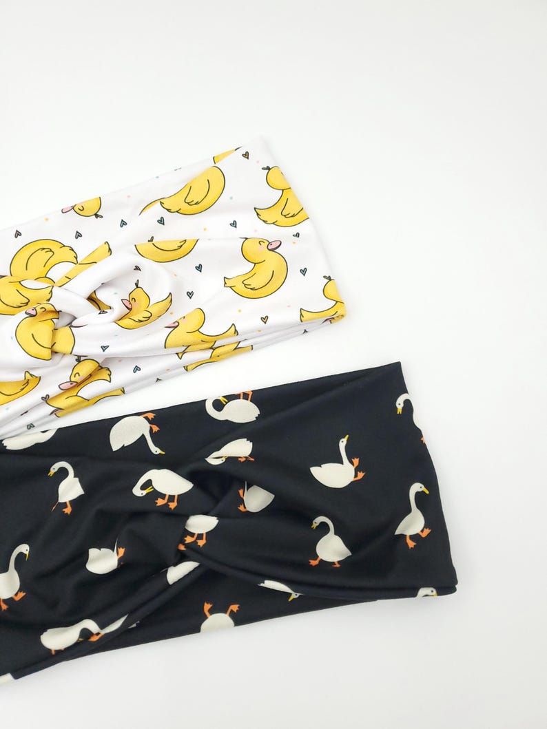 May include: Two headbands with a twist design. One is white with yellow duck illustrations and small hearts. The other is black with white goose illustrations. Both are made of soft fabric.