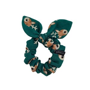 May include: A dark green fabric hair scrunchie featuring a repeating pattern of tan and white deer heads with antlers. The scrunchie includes a tied bow detail at the top and a small black rectangular fabric tag with the white text altness.