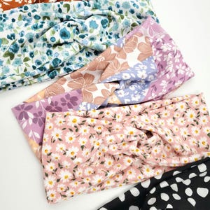 May include: A collection of five fabric headbands featuring a twisted knot design. The patterns include a rust orange floral, a blue and white floral, a lavender and tan floral, a pink daisy print, and a black headband with white polka dots.