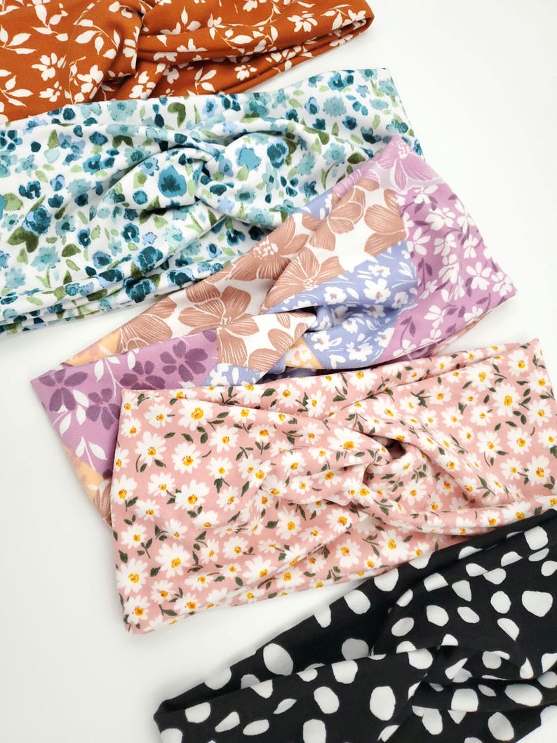 May include: A collection of five fabric headbands featuring a twisted knot design. The patterns include a rust orange floral, a blue and white floral, a lavender and tan floral, a pink daisy print, and a black headband with white polka dots.