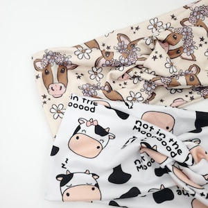 May include: Two headbands with cow-themed patterns. One headband is cream-colored with cow faces wearing flower crowns and floral accents. The other is white with black cow spots and cow faces, with the text "not in the mood".
