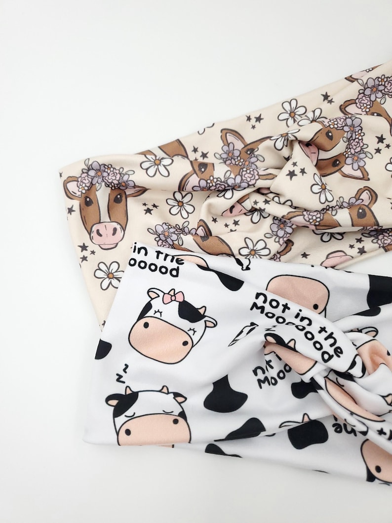 May include: Two headbands with cow-themed patterns. One headband is cream-colored with cow faces wearing flower crowns and floral accents. The other is white with black cow spots and cow faces, with the text "not in the mood".