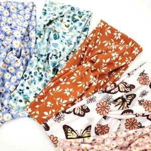 May include: A collection of five fabric headbands featuring various patterns including light blue daisies, blue and green floral prints, a rust orange leaf design, a white background with butterflies and flowers, and a light pink daisy print.