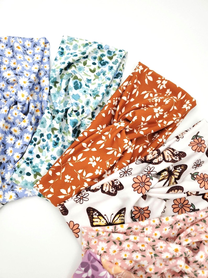 May include: A collection of five fabric headbands featuring various patterns including light blue daisies, blue and green floral prints, a rust orange leaf design, a white background with butterflies and flowers, and a light pink daisy print.