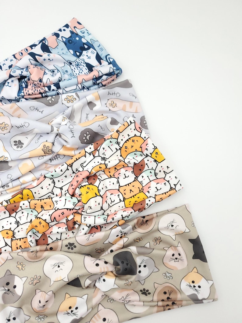 May include: Four colorful headbands with cat patterns. The headbands feature various designs, including cats, fish bones, and paw prints. The colors include blue, gray, white, orange, and tan. These headbands are a fun accessory.