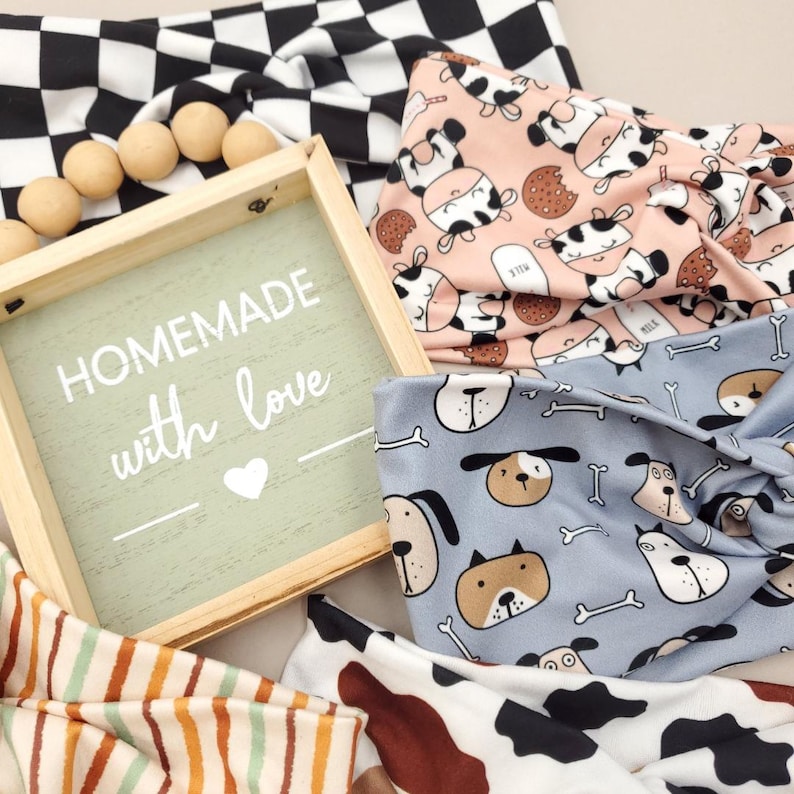May include: Fabric headbands with diverse patterns, including cow print, dog faces, and a black and white checkerboard design. A wooden sign reads "Homemade with love."